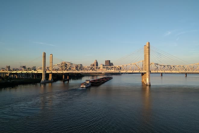 An empty coal barge passes under a brutalist suspension bridge made of white steel and concrete pylons. In the distance, downtown Louisville.