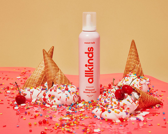 Photo of a light pink shower foam dispenser with the word “allkinds” laid out vertically. Next to the dispenser, four vanilla ice cream cones with rainbow sprinkles sit upside-down, melting, with bright red cherries scattered on top.