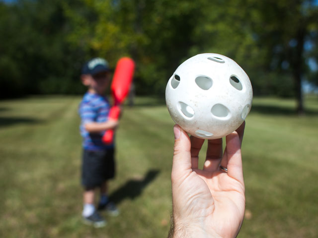 A kid holding a thick red plastic Wiffle Ball bat and a pitcher about to throw a white plastic Wiffle Ball