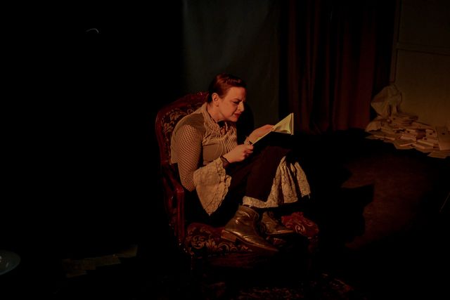 Glow of a lamp on Jane (Lindsey Pierce) reading,
curled up in an arm chair.
