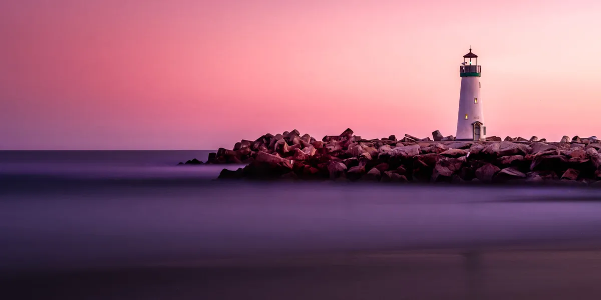 A lighthouse on a rocky shore, with purple water reflecting a rose sunset sky.