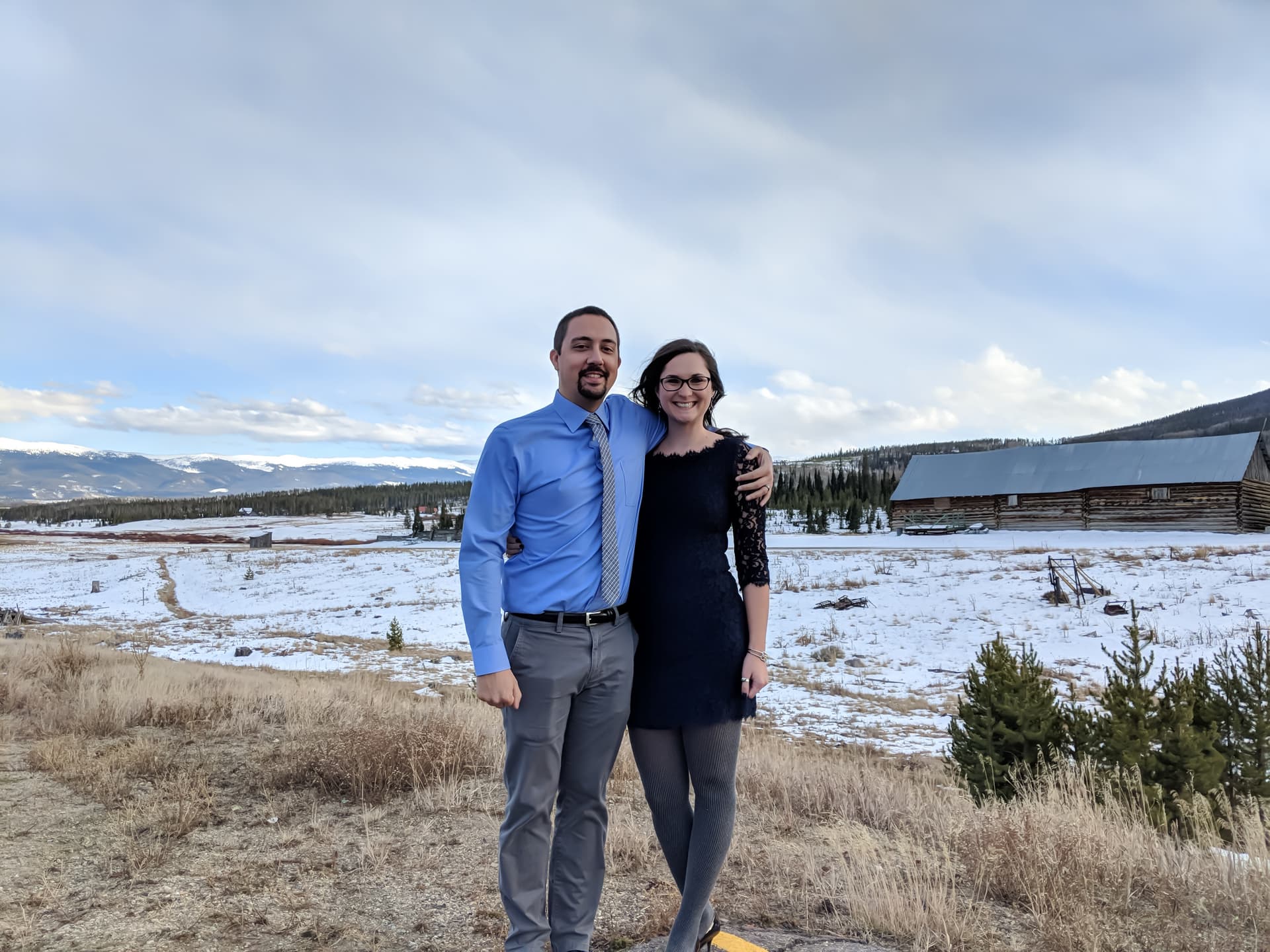 A man and woman pose outside together. Behind them is a snowy high country ranch, and in the distance the Rocky Mountains.
