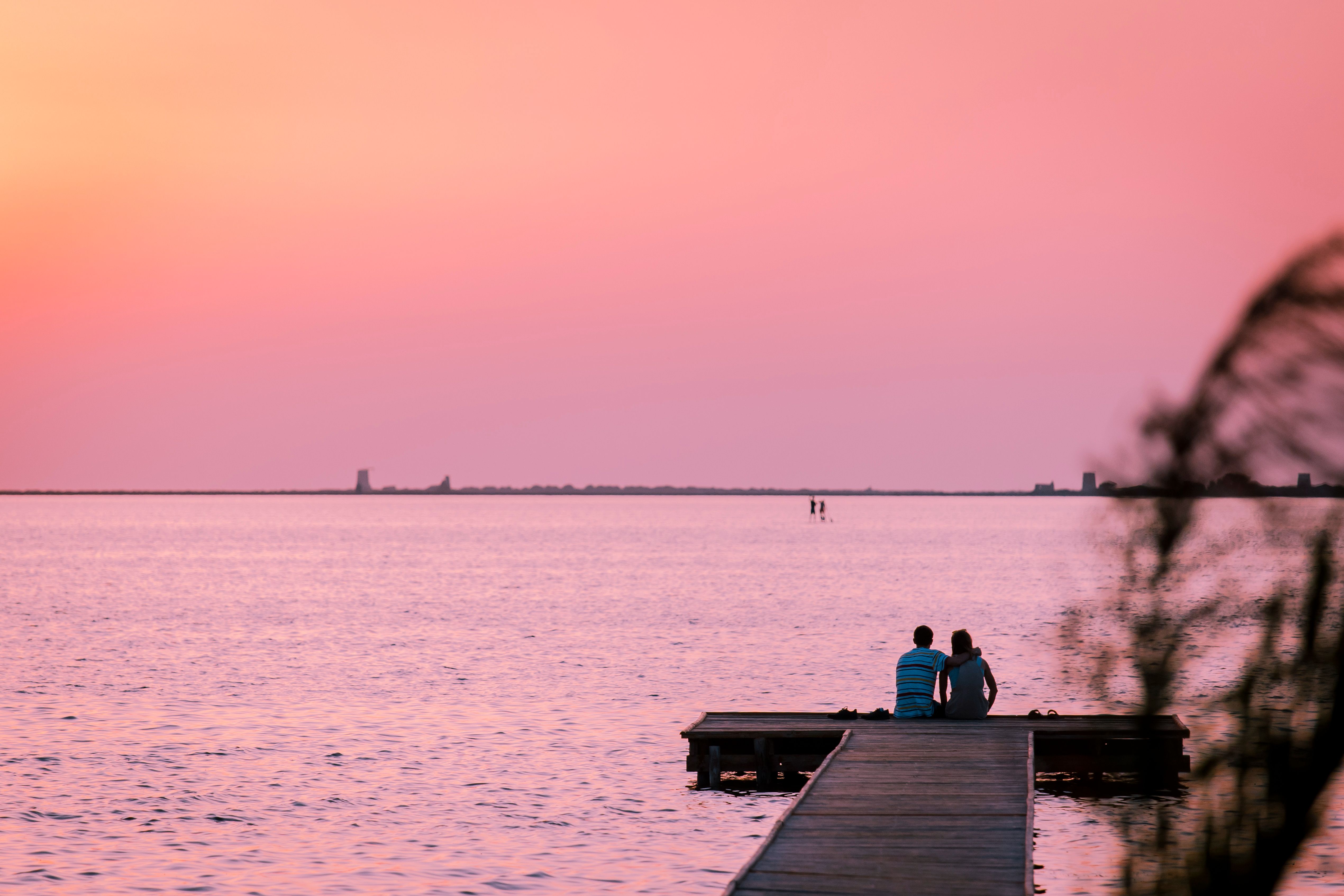 a couple sitting at the edge of a dock at sunset