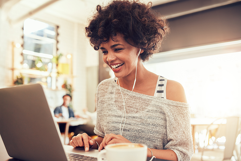 A woman using headphones, smiling at a laptop screen while she types something