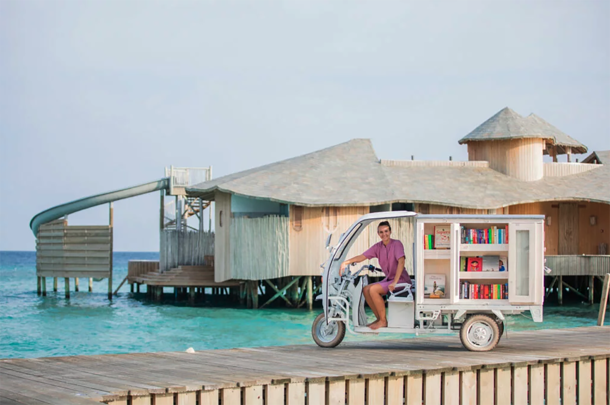 grass huts and a boardwalk over the turquoise blue water in the maldives with a tuktuk on the boardwalk filled with books