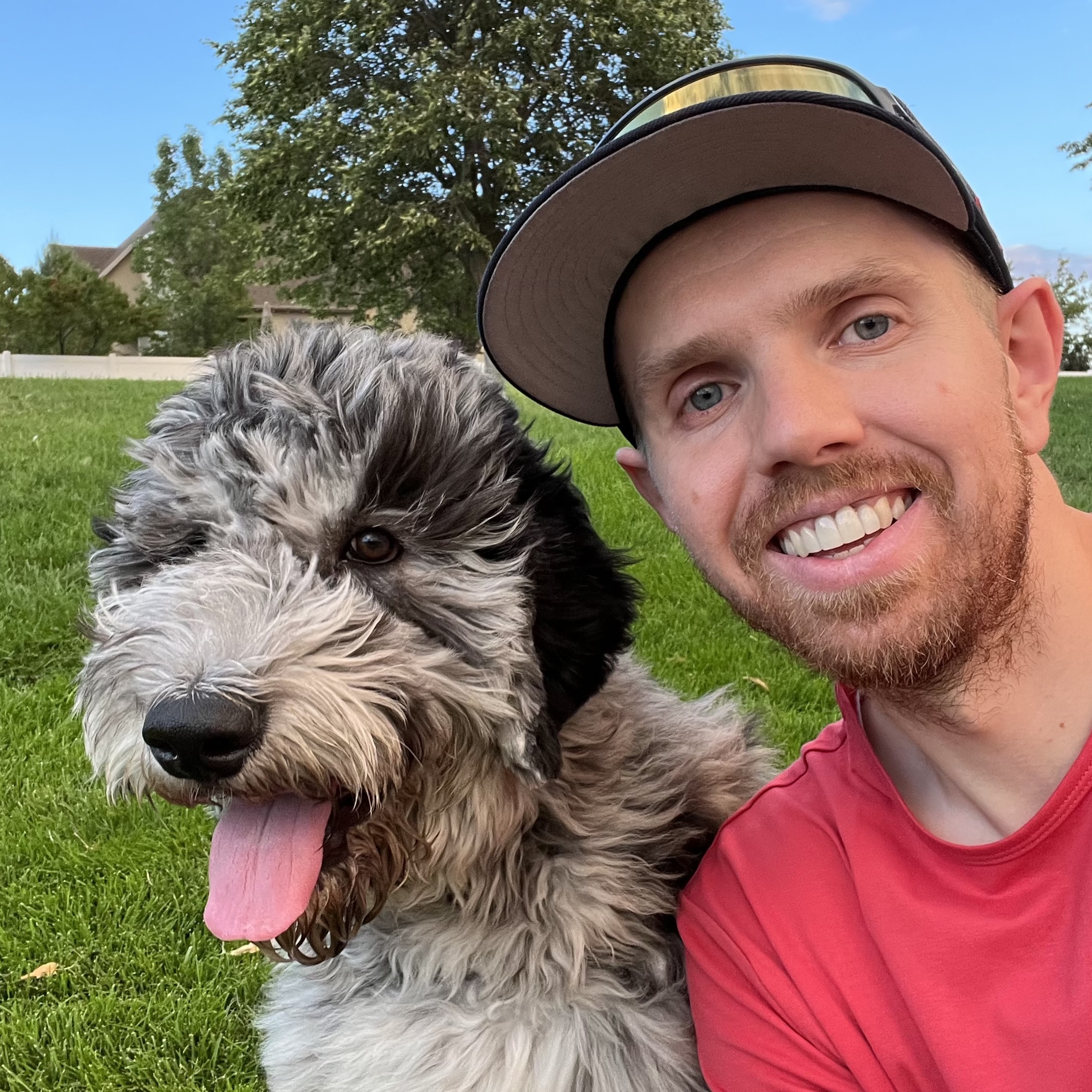 A selfie of my dog Dutch and I on a sunny, windy day with greenery in the background with his tongue hanging out of his mouth