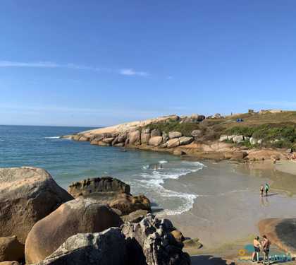 Foto com vista das pedras e faixa de area ao fundo, a esquerda o mar em tom esverdeado.