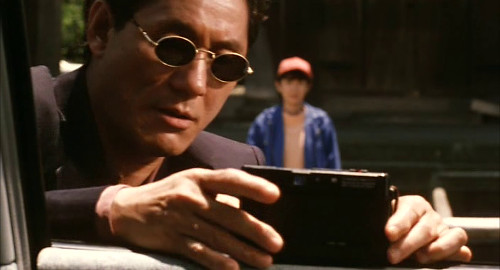 A screenshot from Takeshi Kitano's film 'Hana-bi' of his character in sunglasses setting up a camera for his wife waiting in the background.