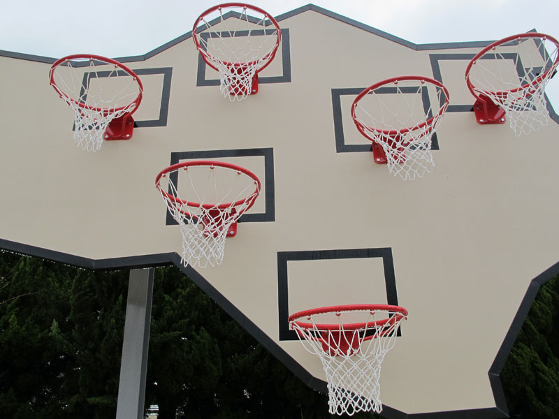 a large, angular, irregularly shaped backboard sits on two poles to make space for six basketball hoops, staggered at different heights