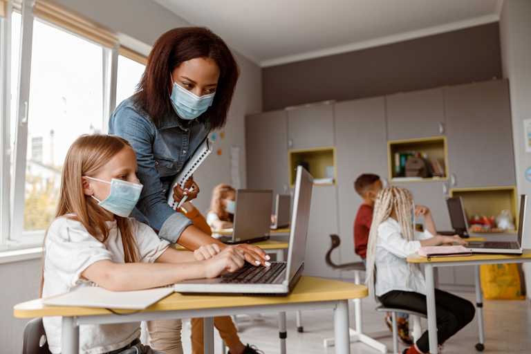 Everyone wearing COVID facemasks, an African American 30-ish teacher helps a young white female student on her computerin an elementary school classroom with two other students in the background.