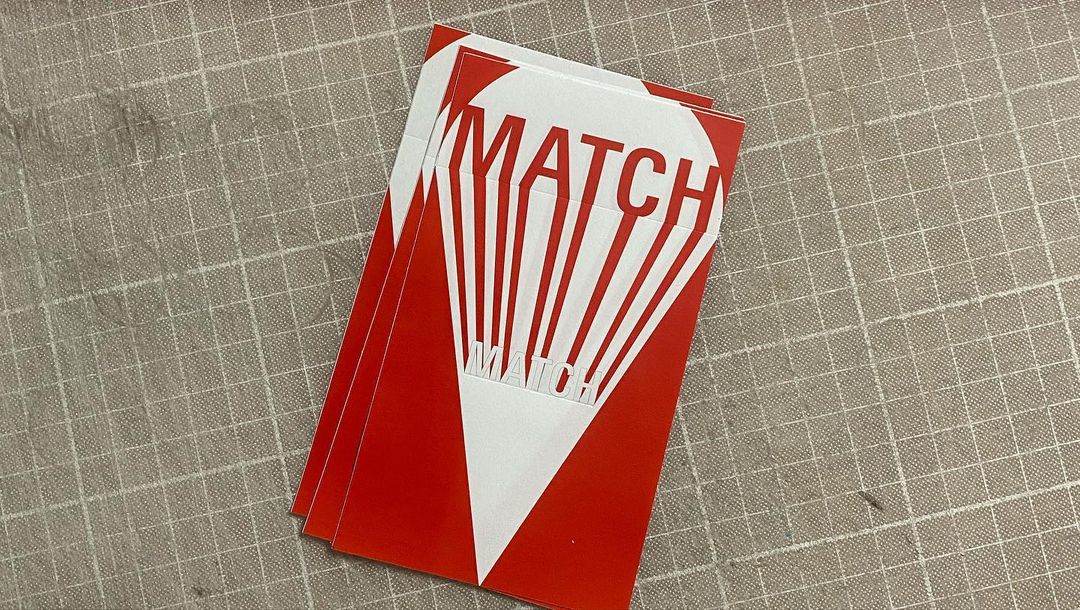 A stack of paper with a red-ink illustration showing the word MATCH in what appears to be the long beam of a flashlight.