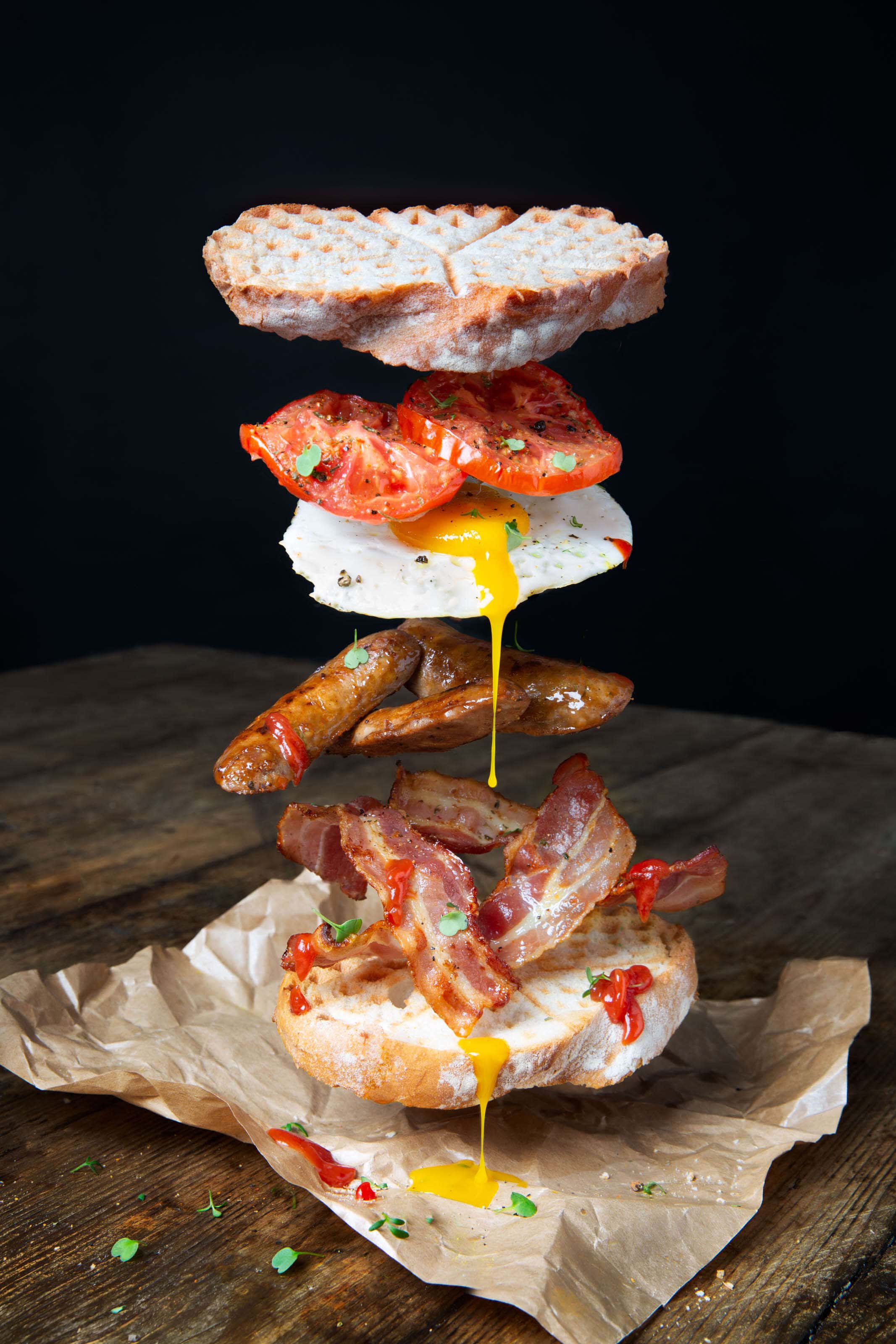 an egg, bacon and sausage sandwich with tomatoes with all ingredients floating above the other. Floating food.