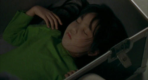 A screenshot from the horrror film 'Seance' of a young girl wearing a green dress lying motionless in a box.