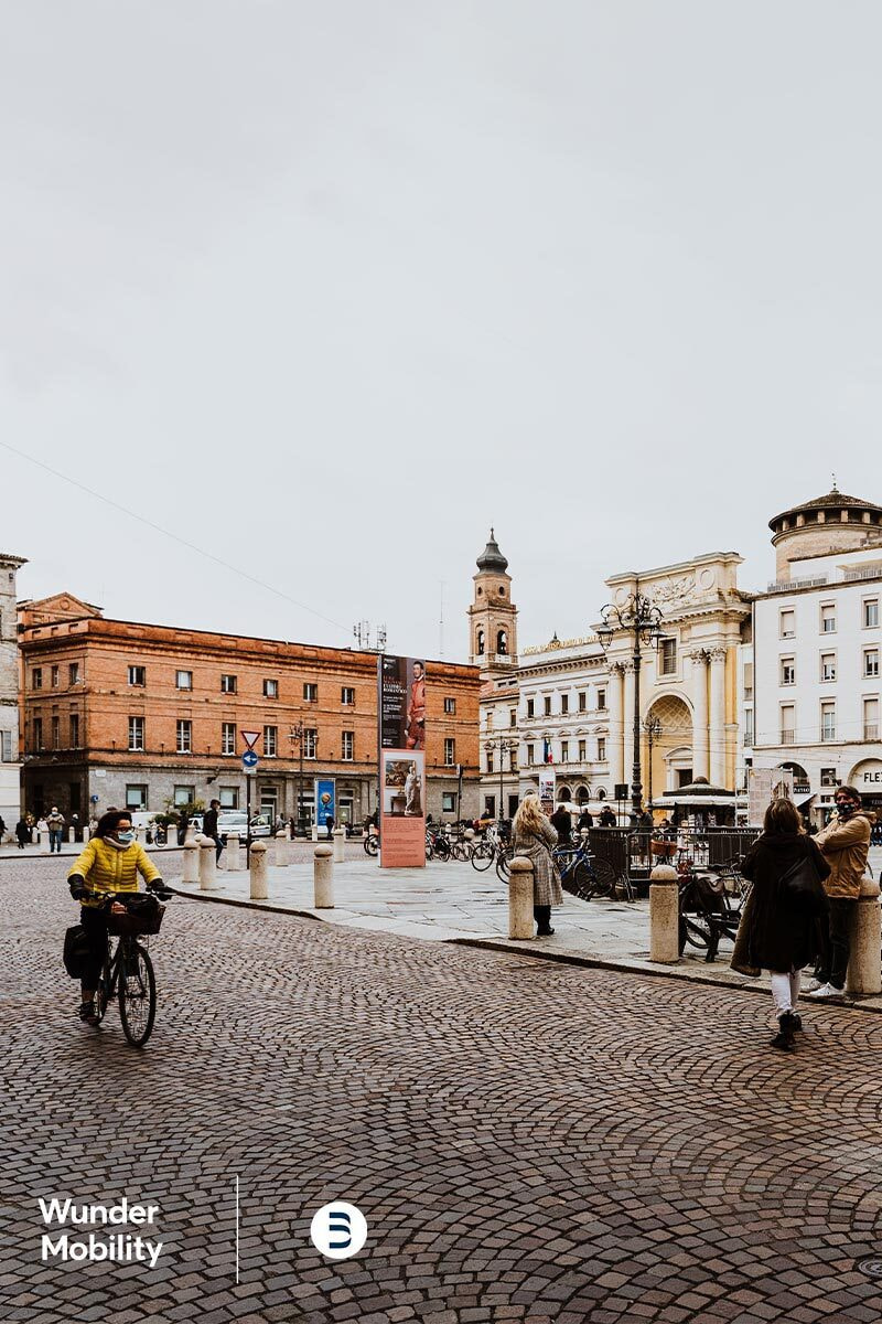 A renaissance styled city square on a busy day with a caucasian person biking across in a yellow raincoat.