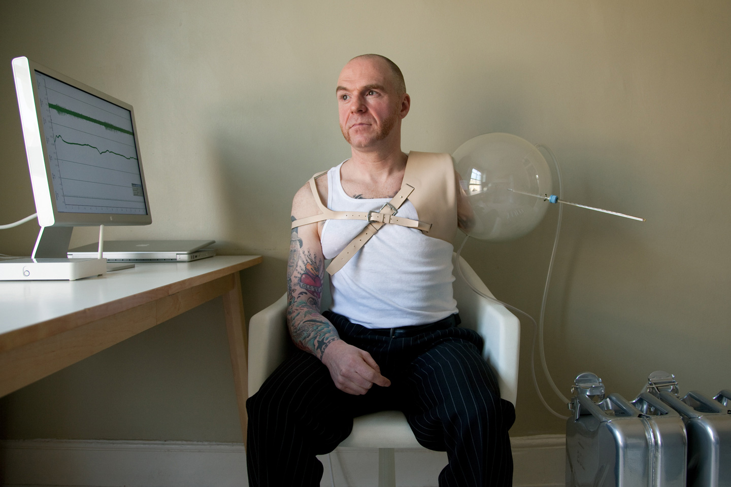 A man sits in a chair; he has one biological arm, and one arm fitted with a balloon-and-straw prosthetic, attached to CPUs. A computer monitor, close by, provides a read-out of data.