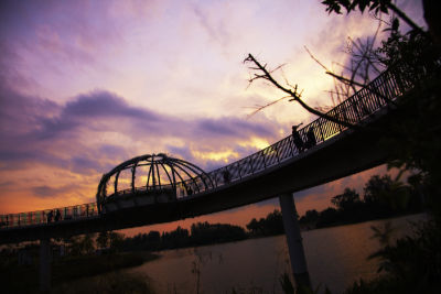 The Jewel Bridge photographed as the sun is setting. The pedestrian bridge pictured over the waterway consists of a dome shaped rest area. A few individuals can be seen on the bridge.