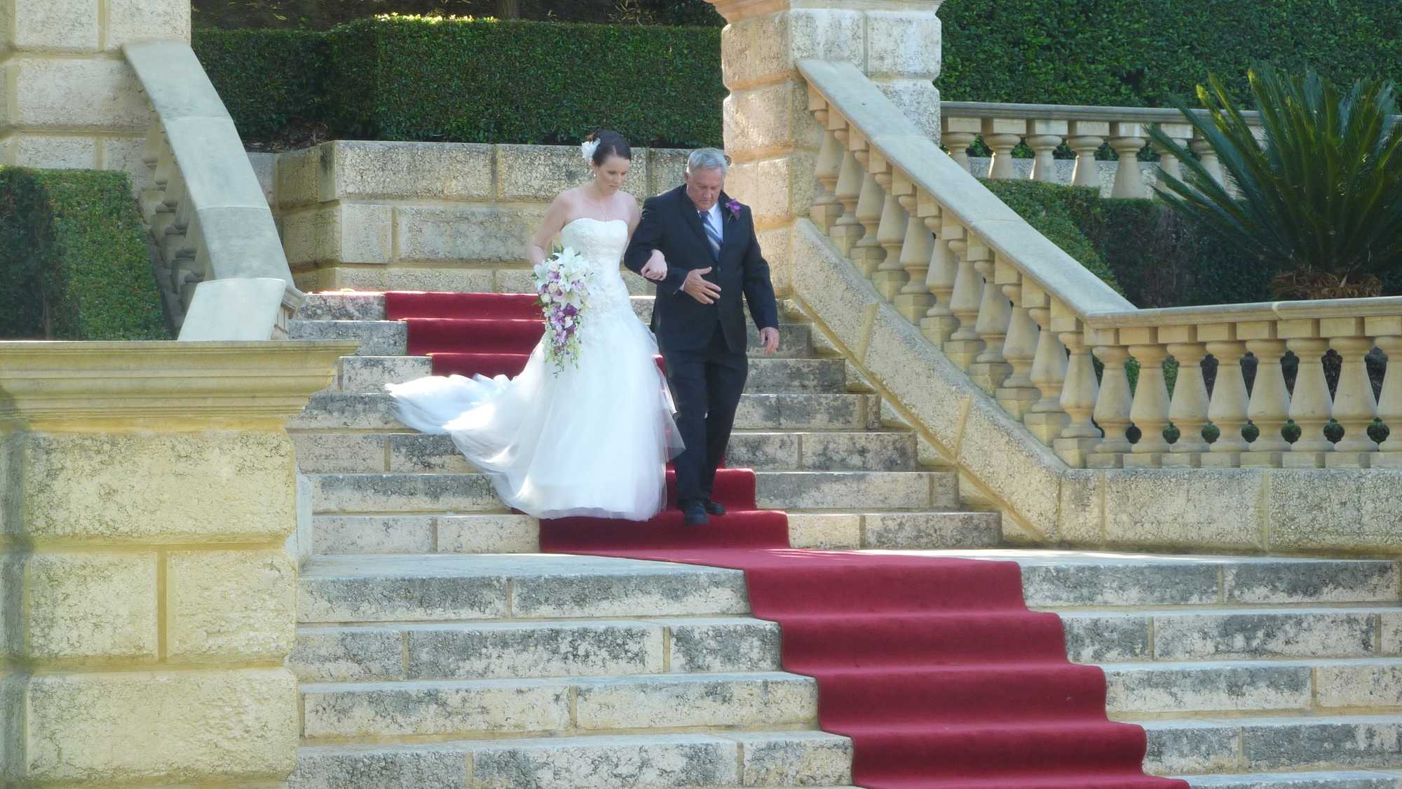 Photo of a bride being walked down a large set of sandstone steps by her father, walking down the aisle lined with a long red carpet. It's outdoors and there are hedges and greenery all around.
