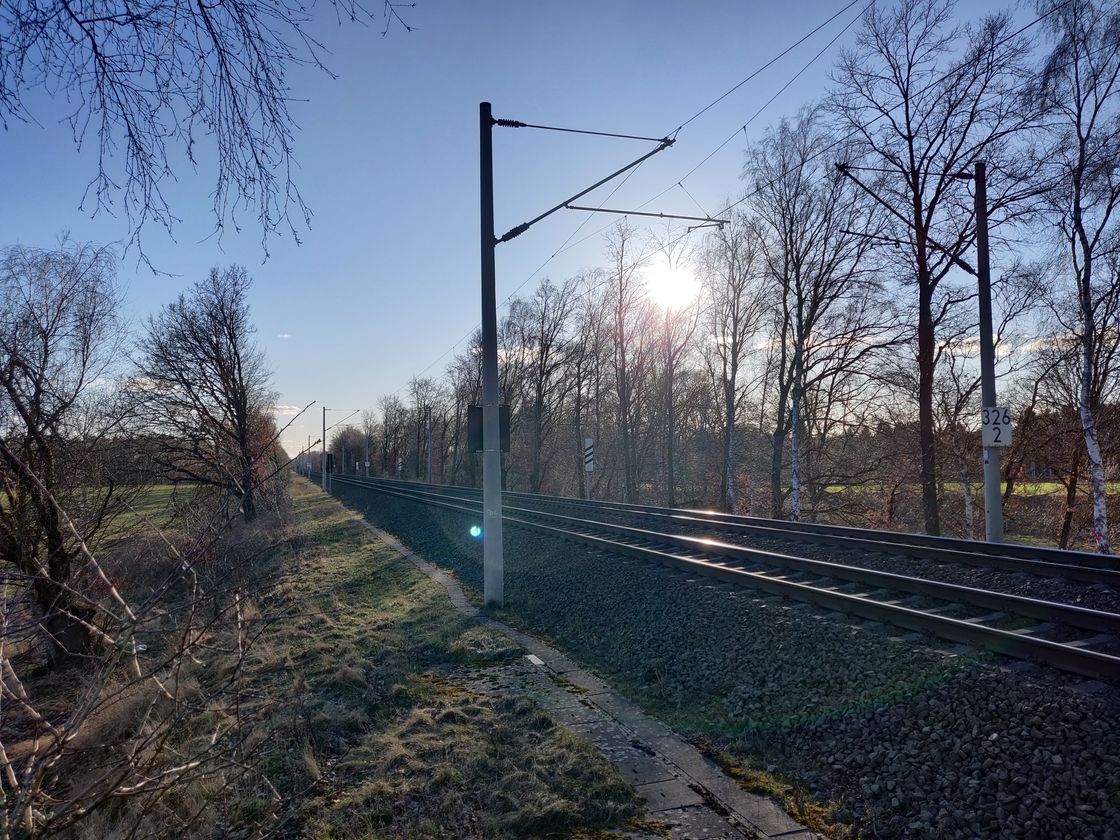 Rail tracks run from the foreground on the right to the horizon on the left. The sun peeks through a line of trees during golden hour just before setting.