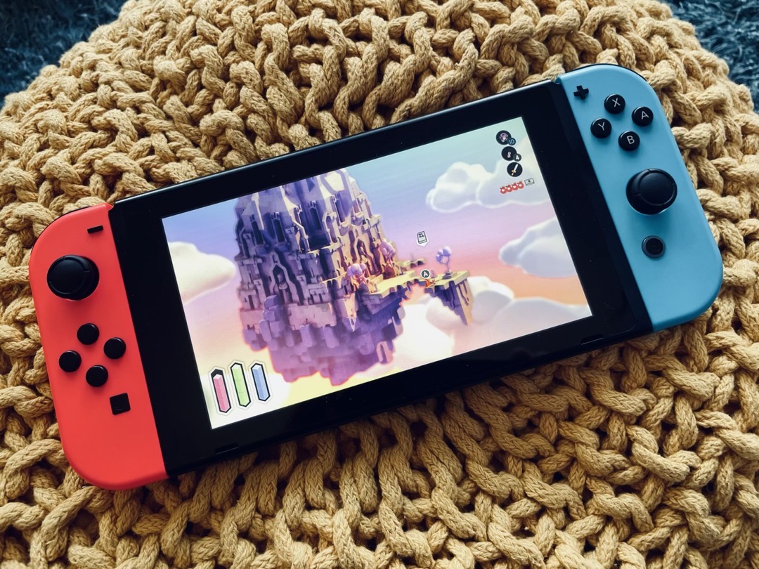 Nintendo Switch in handheld mode, showing a video game in progress. Isometric view with a pastel color palette. A floating castle in the sky. The camera is far from the player, who is barely seen. The graphical user interface show items typical of an action-adventure game, meters for stamina, magic power, etc.