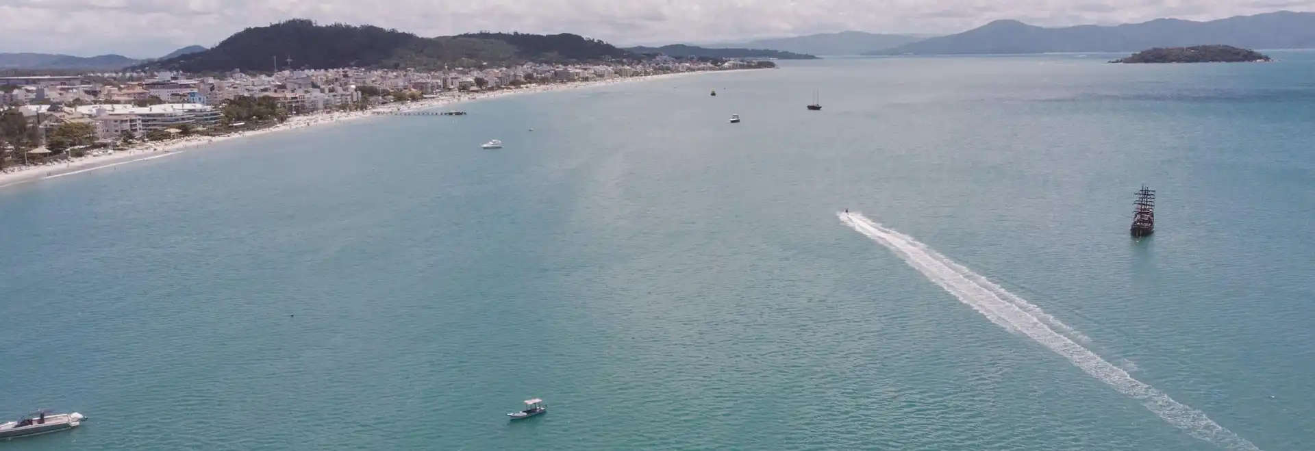 Foto aerea do mar de Canasvieiras com alguns barcos navegando e faixa de areia bem ao fundo.