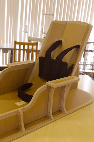 A floor sitter of cardboard with black neoprene straps for support, arm rests and a wide base.