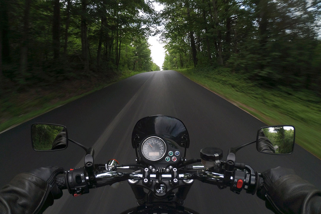 Riding a motorcycle from my point of view, my hands, handlebars, and speedometer in the foreground, going down a freshly paved road surrounded by trees.