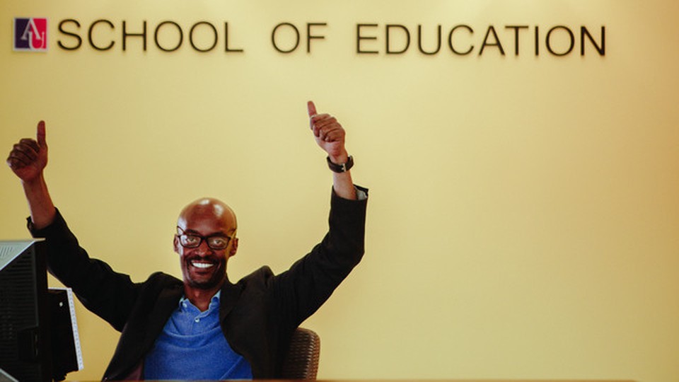 A man seated in front of a wall written School of Education. He has with both hands raised and is smiling.