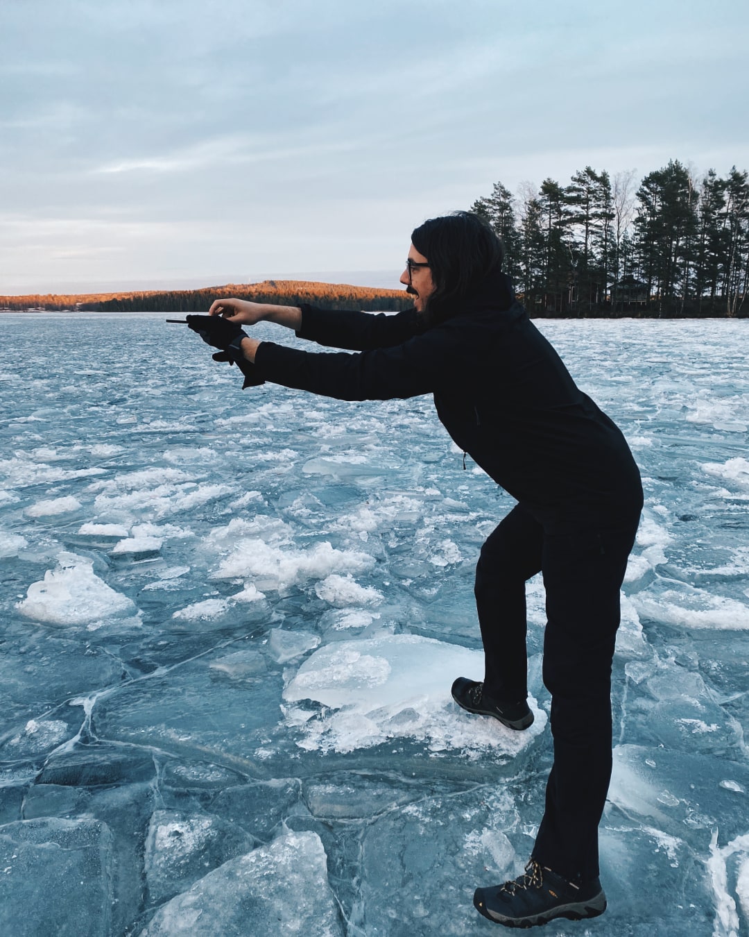 A man is standing on a frozen lake. He's holding a smartphone far away from his body in an awkward position. He's probably about to take a picture.
