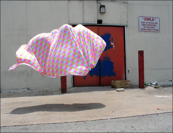 A pink and blue printed sheet of fabric hangs suspended in the air, mountainous in its shape. In the background, a security sign on the wall reads: SMILE, your activities are being recorded...