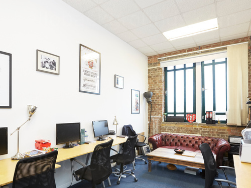 Small office workspace lined with wooden computer desks with chairs and featuring red chesterfield sofa in front of window Zoom background
