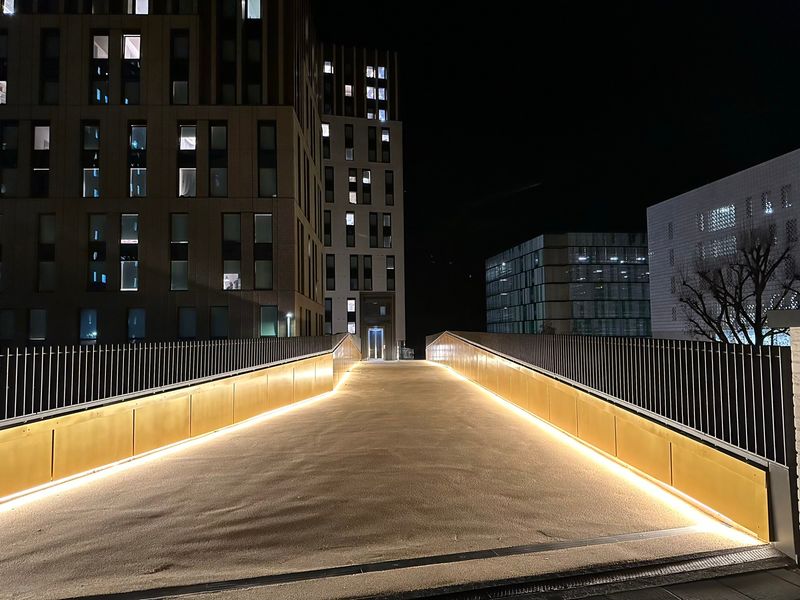 Taken at night. The bridge has downlighting on either side of the path, showing the light brown colour and slightly rough texture (for grip). On the other side of the bride is the entrance to an elevator down to street level. Some lights are on in the high rise university residences behind.