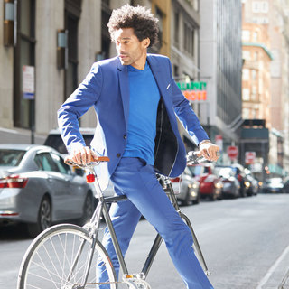 A man in a blue suit crosses a city street with his bike.