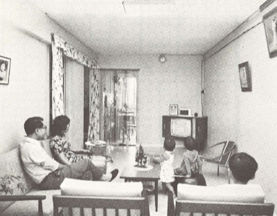 A black and white photo of a family of five watches television in the living room of their three-room HDB flat.