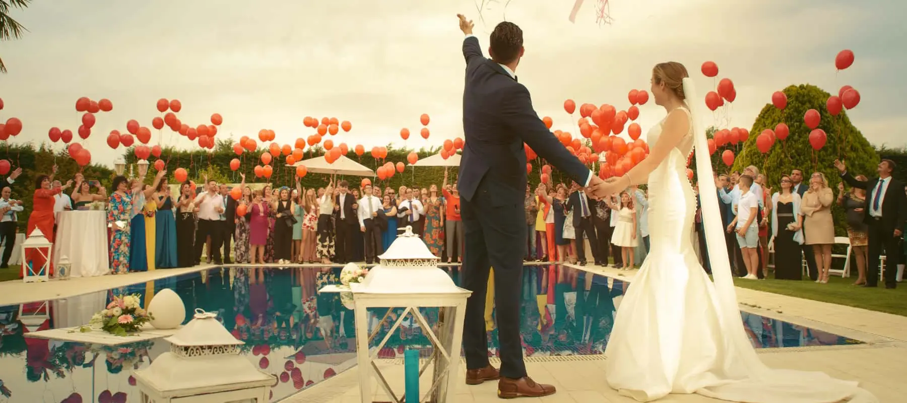 Um casal vestido com trajes de casamento, de mãos dadas, segurando balões de festa e acenando para seus convidados segurando balões vermelhos do outro lado de uma piscina ao ar livre