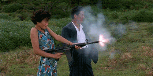An animated gif of a scene from Takeshi Kitano's film 'Sonatine' of a woman shooting an automatic rifle with a yakuza man standing next to her.