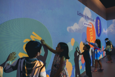A photo of the Words That Move room. Children are pressing their hands onto a wall projection. The words, 'Listen to the sound' is displayed on the image.