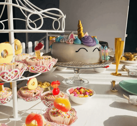 Uma mesa de lanche colorida com cupcakes, gomas, flutes douradas e, no centro da mesa, um bolo com a forma de um unicórnio