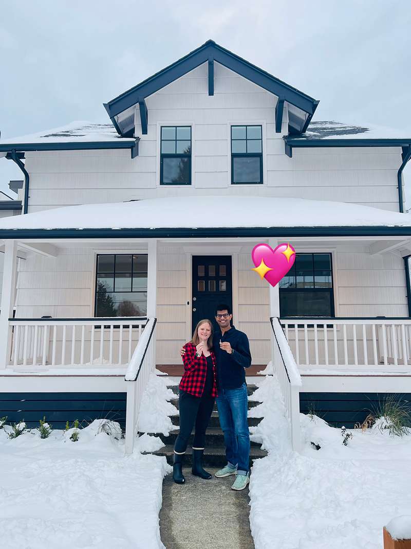 My boyfriend and I holding up keys in front of a cream-colored farm-style house with black and white trim. There is heavy snow on the roof and small lawn in front of the house.