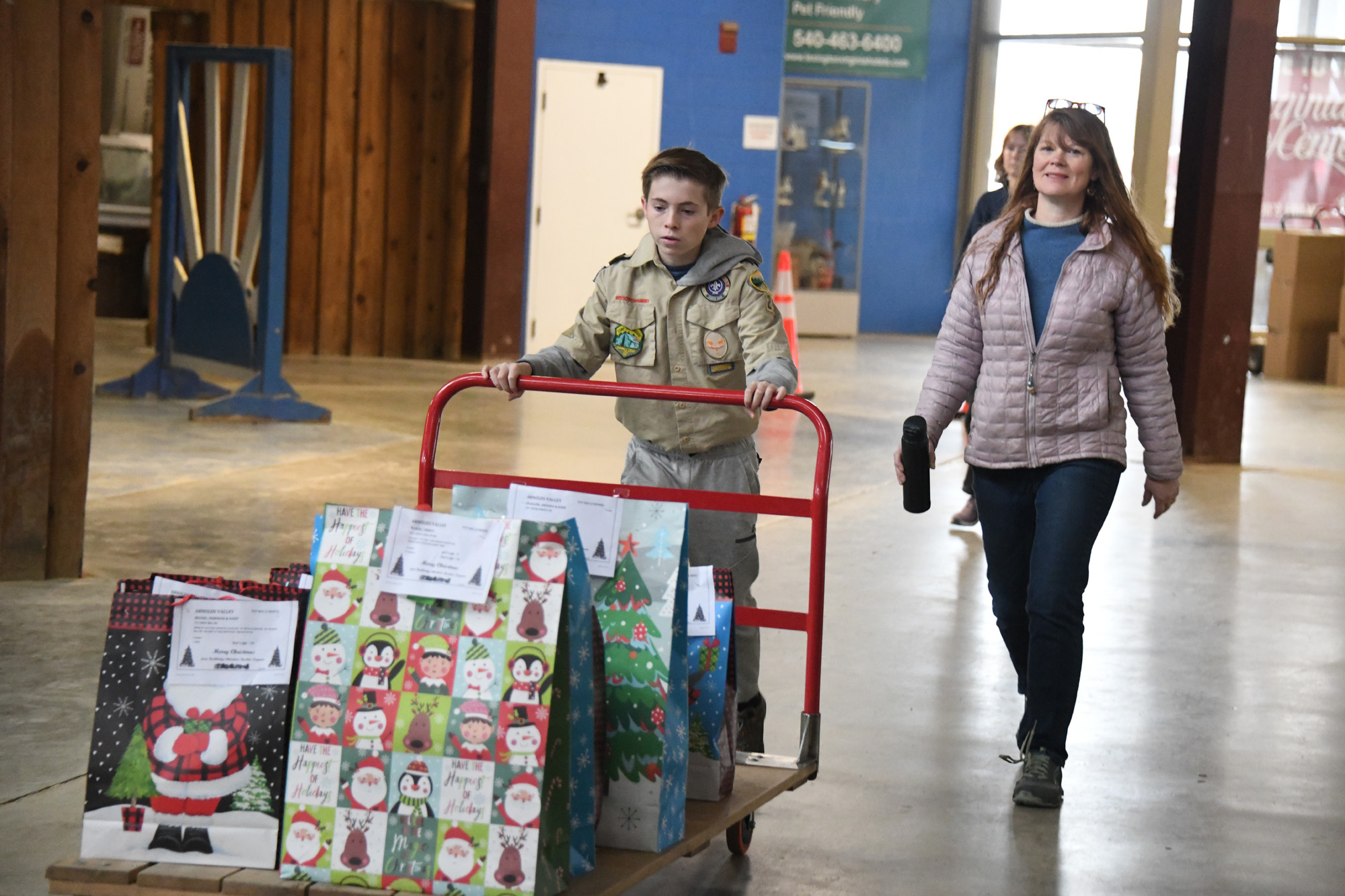 Tina Miller walking behind a boy scout pushing a cart loaded with brightly colored Christmas gift bags full of toys.