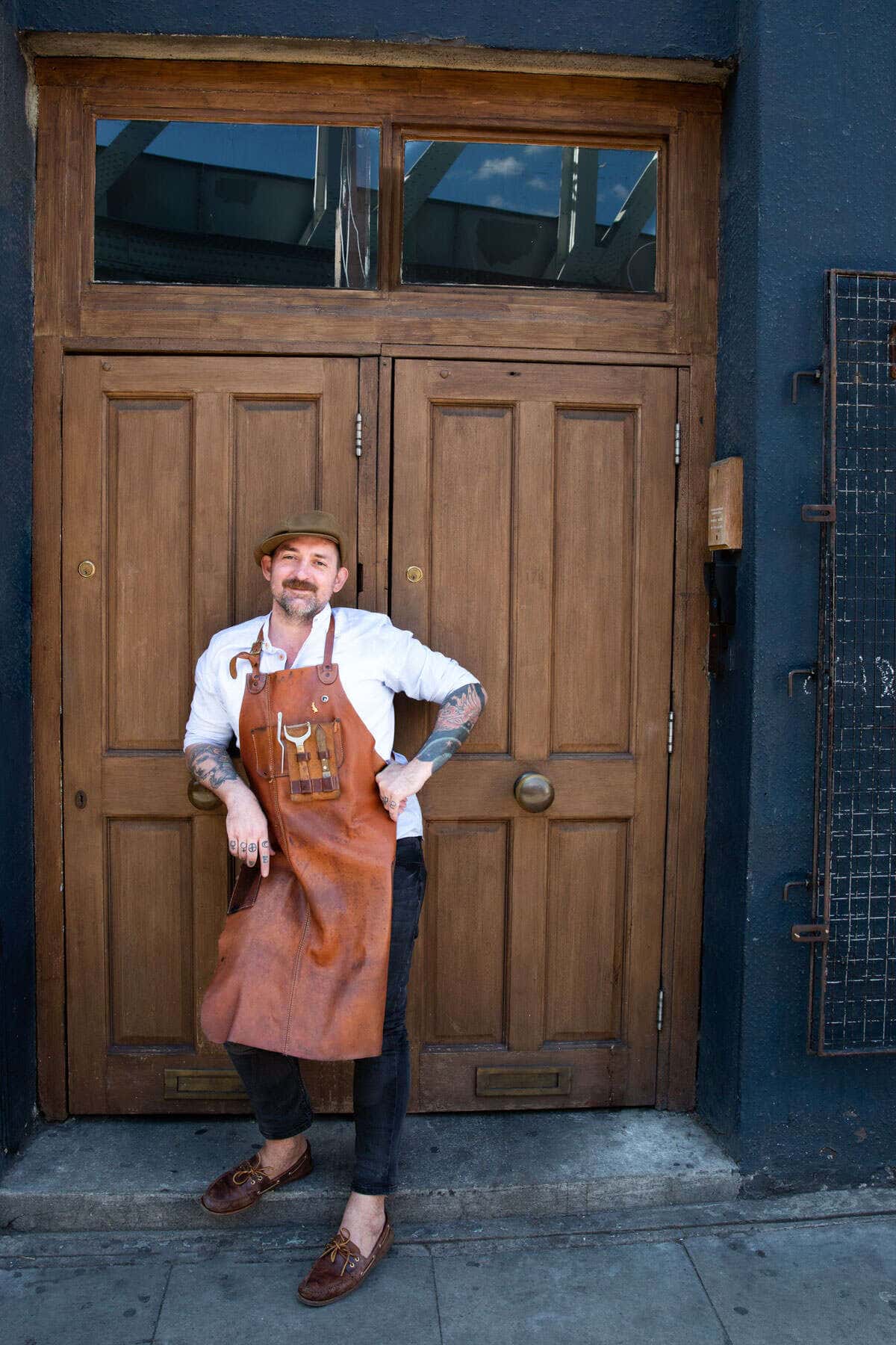 A portrait of a bartender in his apron stood in front of some large wooden doors.