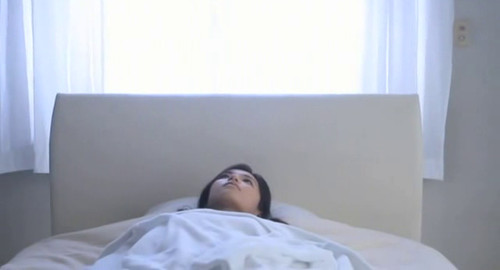 A screenshot from the movie 'Nude' of a young woman lying in bed under the covers looks up.