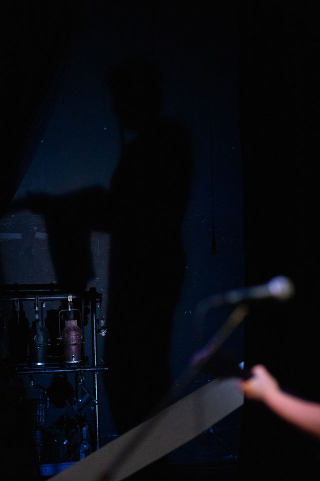 Josie's mic and hand in the foreground
but the focus is Miriam's shadow cast in blue light
on the back wall of the theater
next to a rack of stage lights
