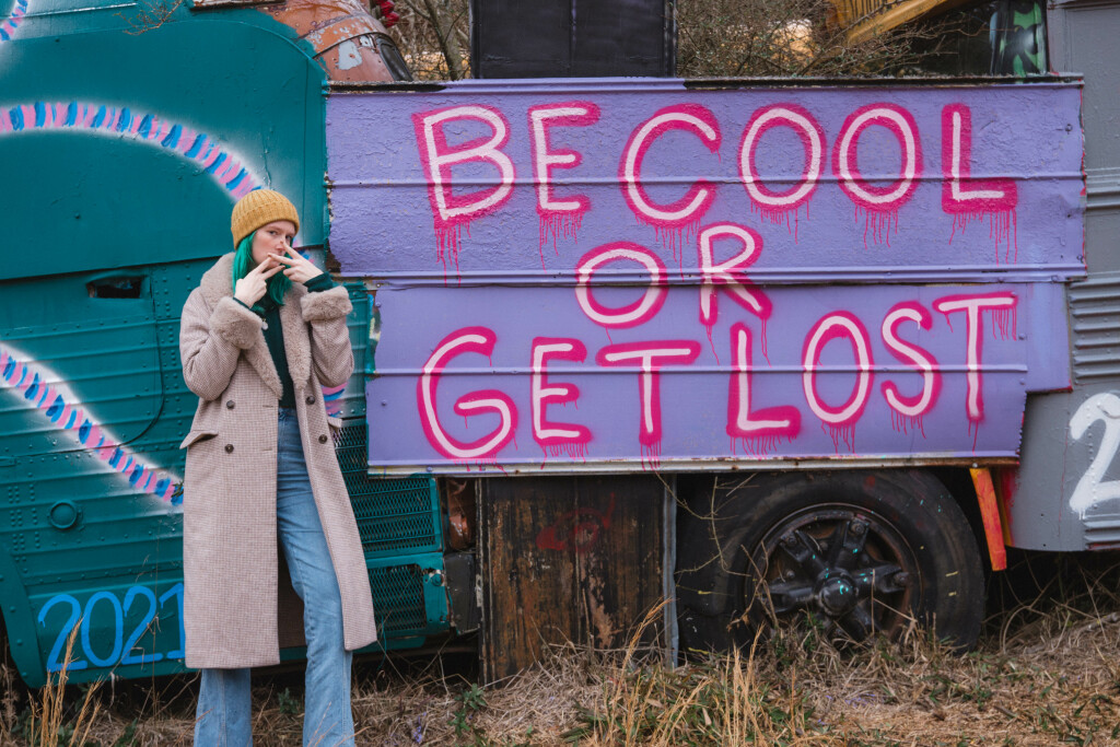 Beth wears a long coat in front of a sign that says Be Cool or Get Lost