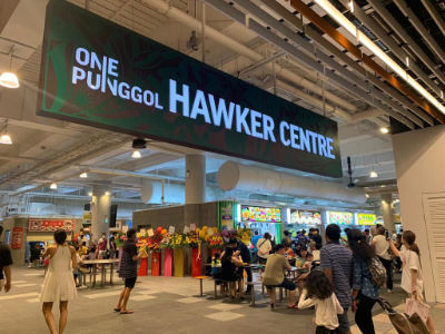 People enter the One Punggol Hawker Centre during its opening in October 2022. A green rectangular signboard hangs above the entrance, reading 'One Punggol Hawker Centre'.