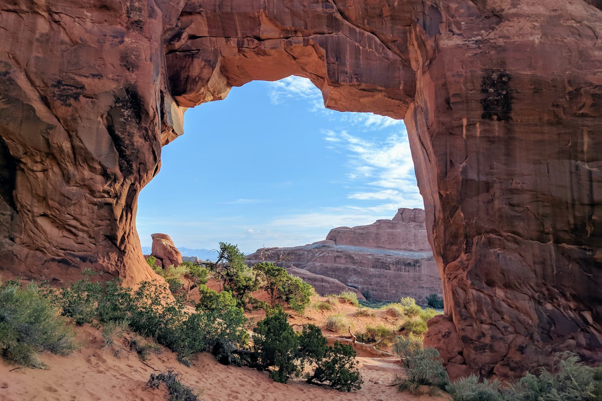 Looking through Pine Tree Arch in Arches National Park. The stone of the arch is exceptionally red, and the ground very sandy. Small desert shrubs grow on either side of the arch.