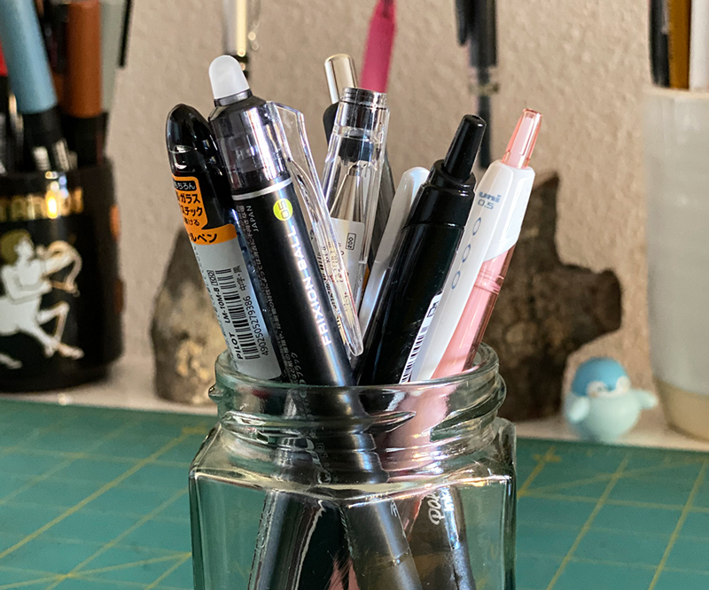 A cup of pens on a desk, with more pens in the background