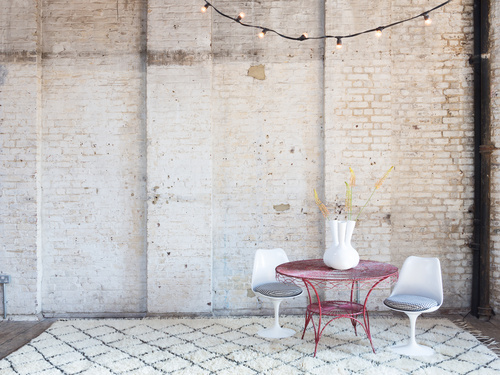 Rustic workspace featuring painted brick wall behind pink metal table and two white modern chairs on white chequered rug  Microsoft Teams background