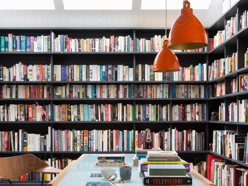 Large black corner bookcase adorned with books behind glass desk with wooden frame and wooden chair Google Meet background