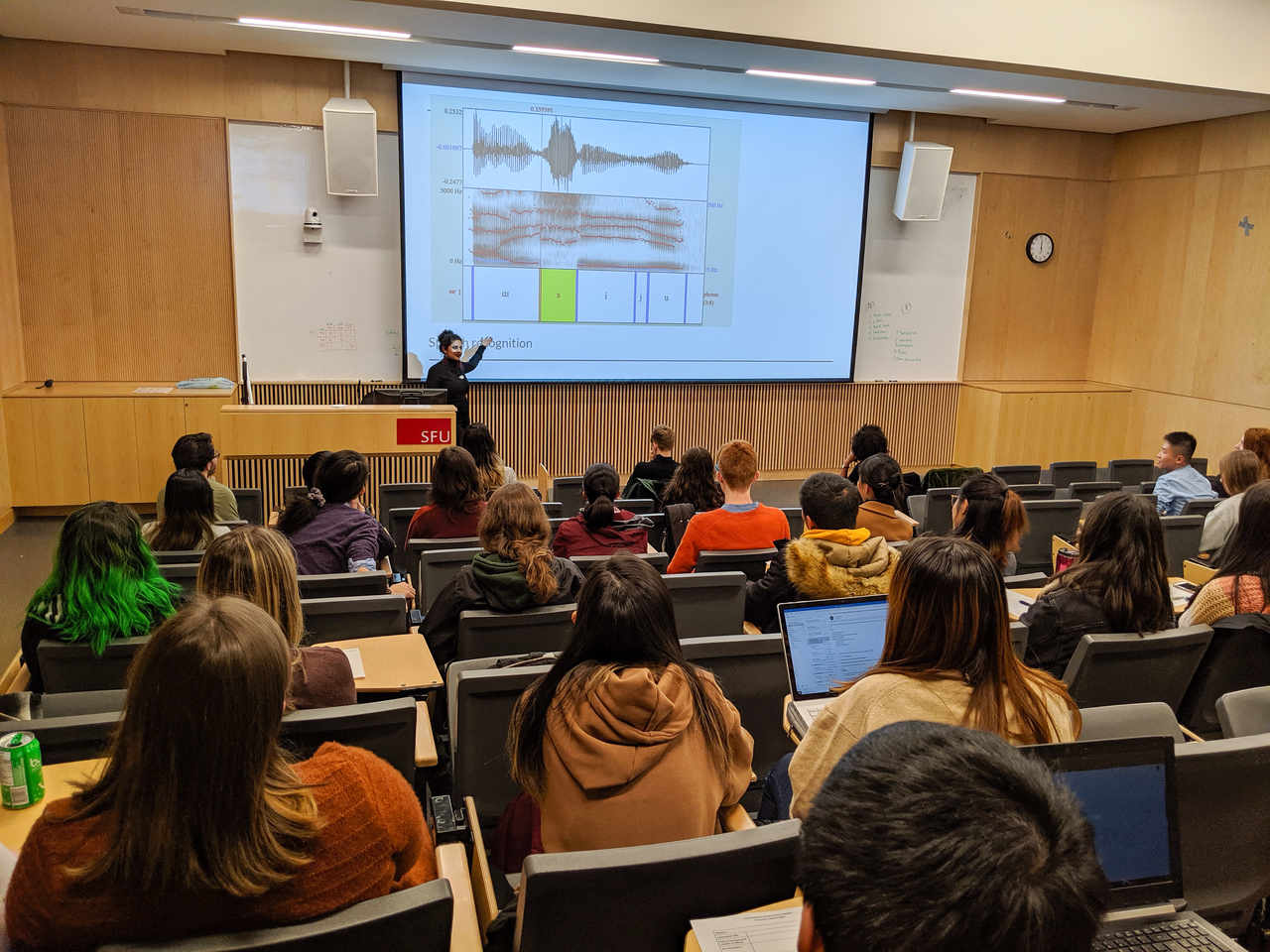 Vasundhara is presenting at a lectern in front of a screen on which a screenshot of the computer program Praat is projected. There are about 30 people listening to xem present and the photo shows the backs of their heads.