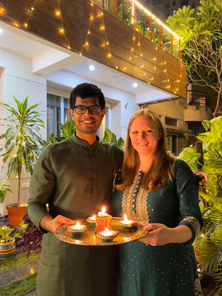 My fiance and I holding a tray of candles in front of house decorated with lights. We are wearing traditional Diwali shirts in tones of dark green.